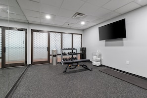 A gym with a bench press, dumbbells, a TV, and a trash can.