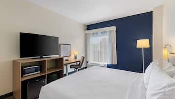 Habitación de hotel con cama, escritorio, televisor, lámpara y ventana con cortinas.