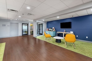 A spacious indoor area with a flat ceiling, a television mounted on the wall, and a seating area with chairs and a small table. - Spark by Hilton Waynesboro (Waynesboro)