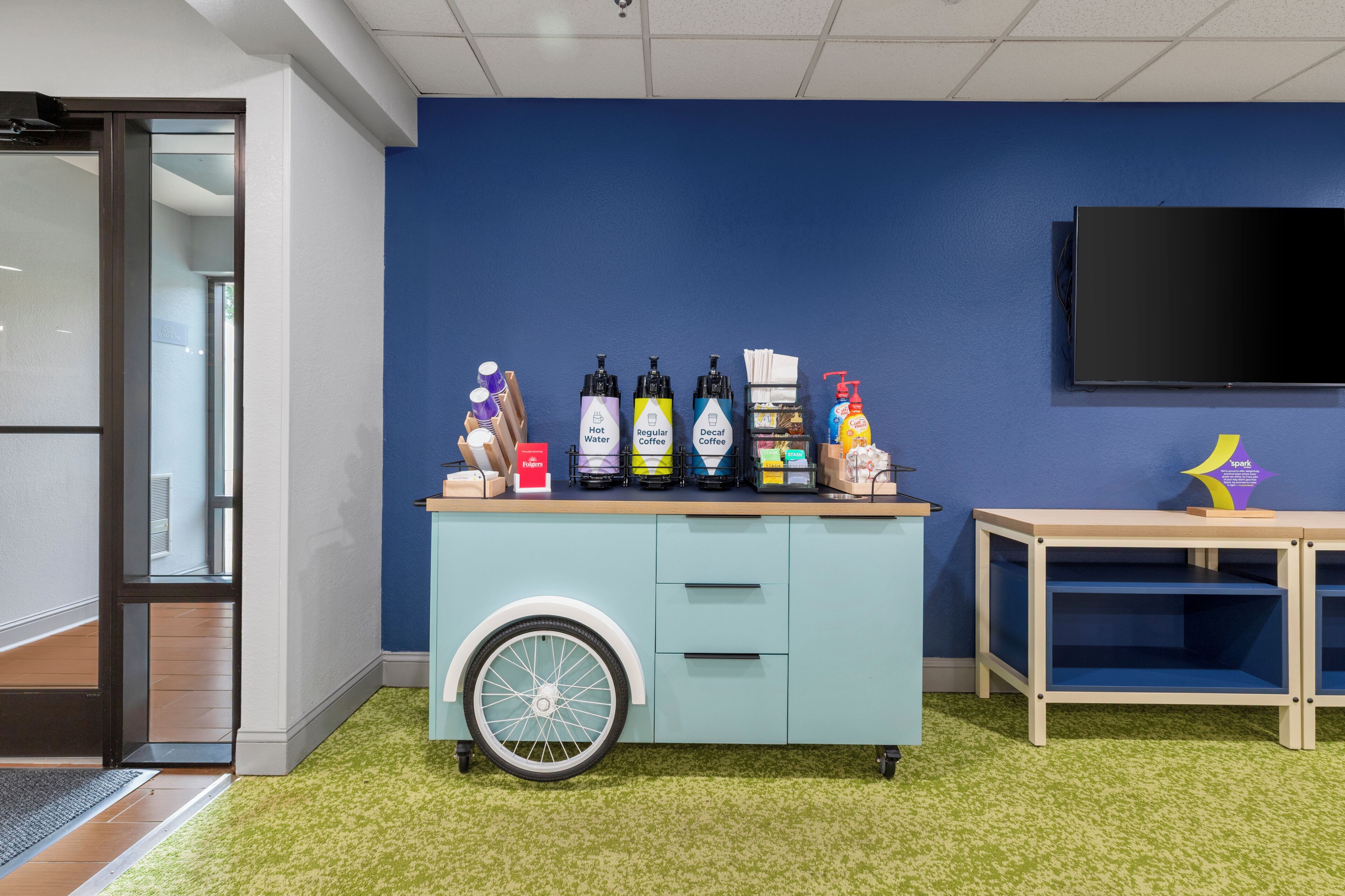 Una habitación con pared azul, un carrito pequeño con varios objetos, un televisor y una alfombra verde.