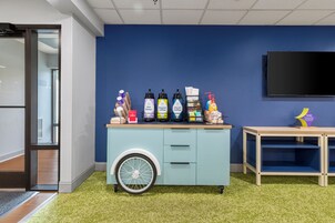 Una habitación con pared azul, un carrito pequeño con varios objetos, un televisor y una alfombra verde.