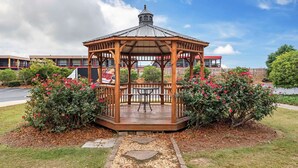 A wooden gazebo with a metal roof, surrounded by flowering bushes and a stone pathway.