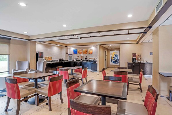 A dining area with multiple tables and chairs, a kitchen in the background, and artwork on the walls. - Comfort Inn Opelika - Auburn (Opelika)