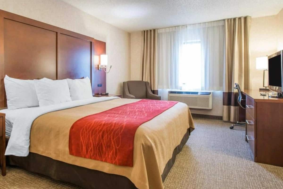 Ein Hotelzimmer mit einem großen Bett, einem Schreibtisch mit Computer, einem Stuhl und einem Fenster mit Vorhängen.