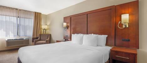 Quarto de hotel com uma cama grande, uma cadeira, mesa de cabeceira com abajur e luminária na parede.