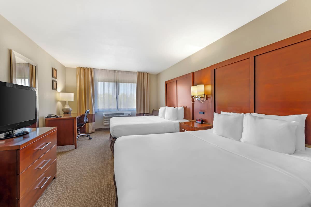 Ein Hotelzimmer mit zwei Betten, einem Fernseher, einem Schreibtisch, einer Lampe und einem Fenster mit Vorhängen.