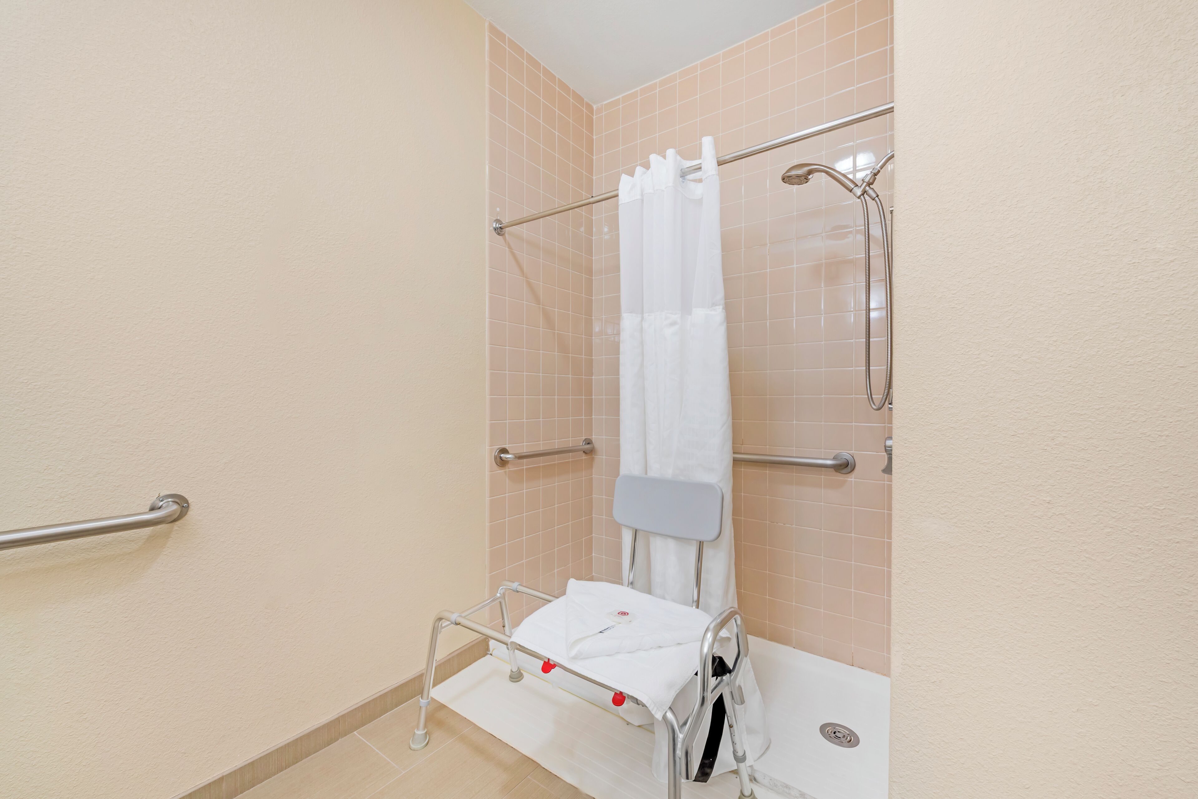 Ein Badezimmer mit Dusche, Haltegriff, einer rollstuhlgerechten Sitzbank und einem weißen Vorhang.