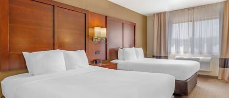 A hotel room with two beds, wooden headboards, a nightstand with a lamp, and a large window with curtains.