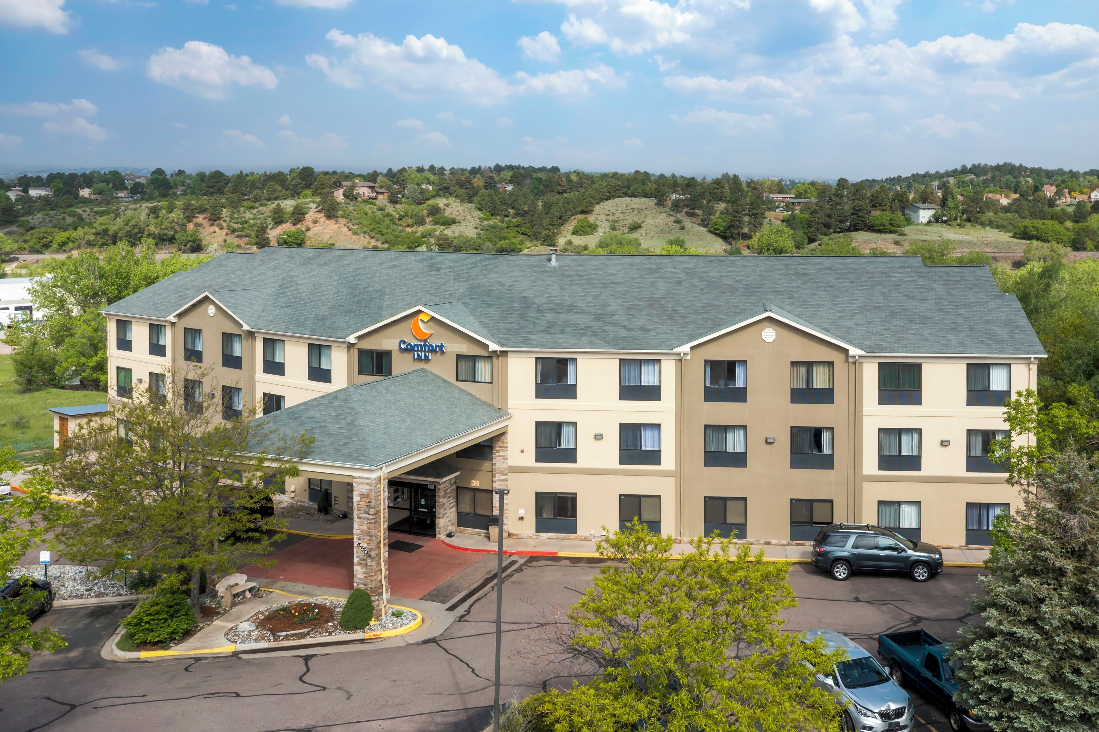 A multi-story hotel with a central entrance, a parking lot, and a hilly landscape in the background.
