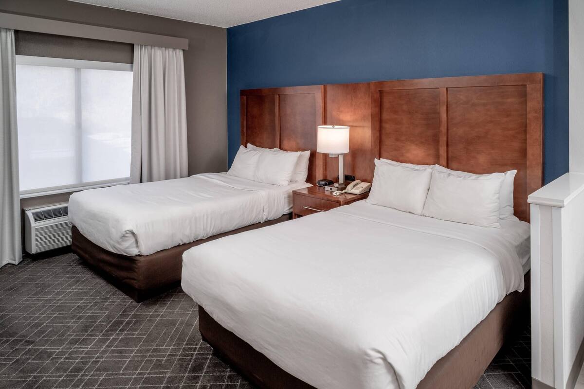 Een hotelkamer met twee bedden, een houten hoofdbord, een nachtkastje met een lamp en een raam met gordijnen.