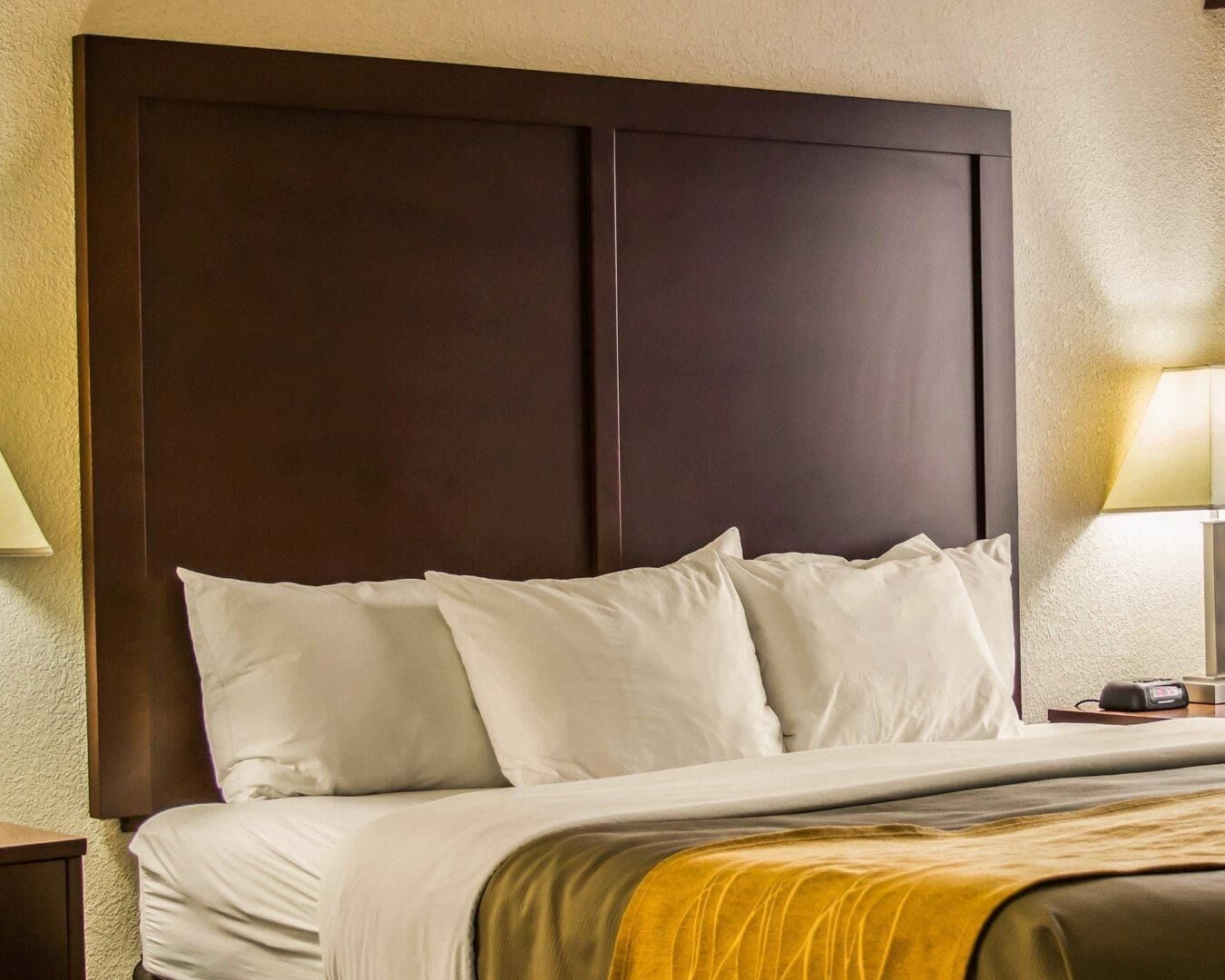 Een hotelkamer met een groot, donker hoofdeinde, een bed met witte lakens en een nachtkastje met een lamp.