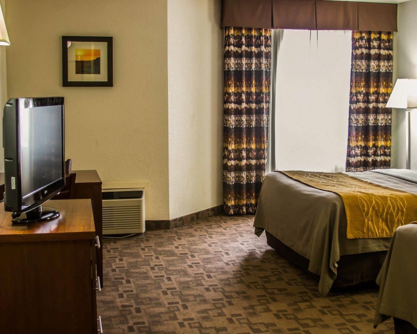A hotel room with a bed, a TV, a desk, and a window with curtains.