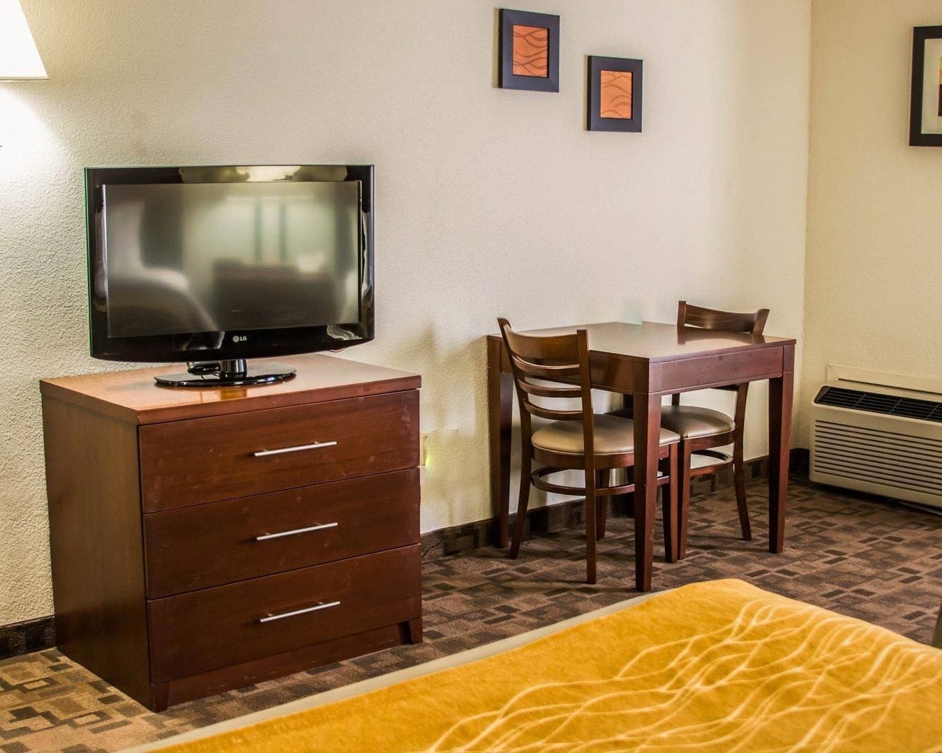 A hotel room with a TV on a wooden cabinet, a dining table with chairs, and a patterned rug.