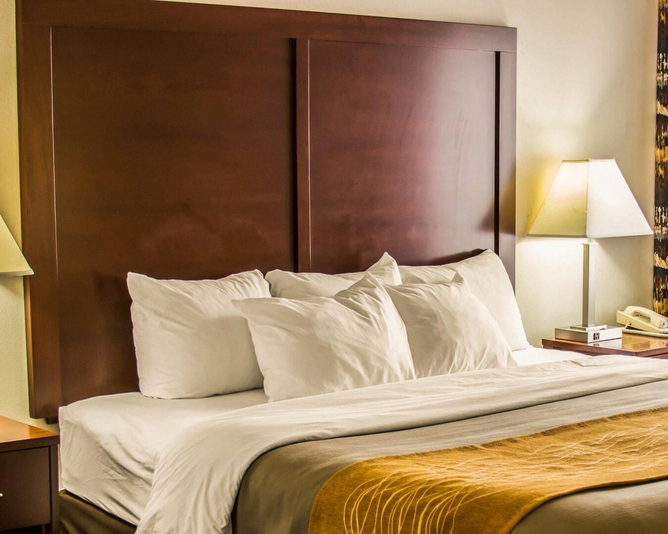 A hotel room with a large wooden headboard, a bed with white linens, a bedside lamp, and a telephone on the nightstand.