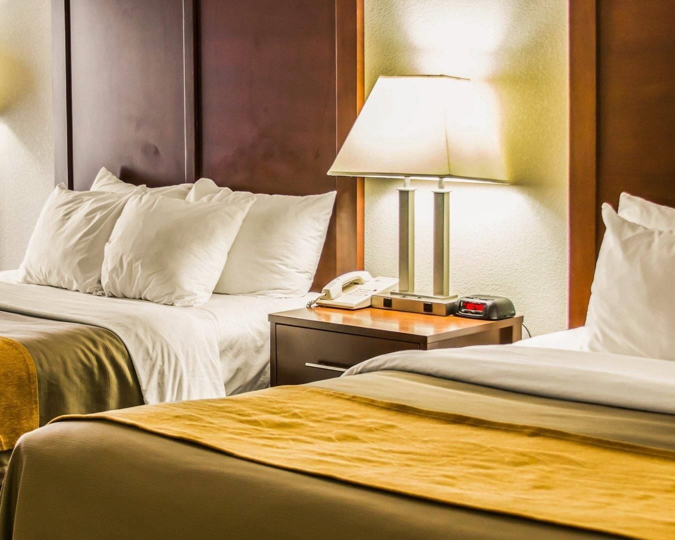 A hotel room with two beds, a bedside table with a lamp, a phone, and a clock.