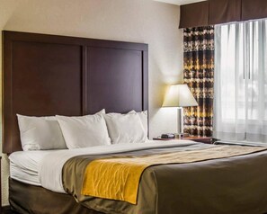 A hotel room with a large bed, a bedside table with a lamp, and a window with curtains. - Comfort Inn Birch Run - Frankenmuth (Birch Run)