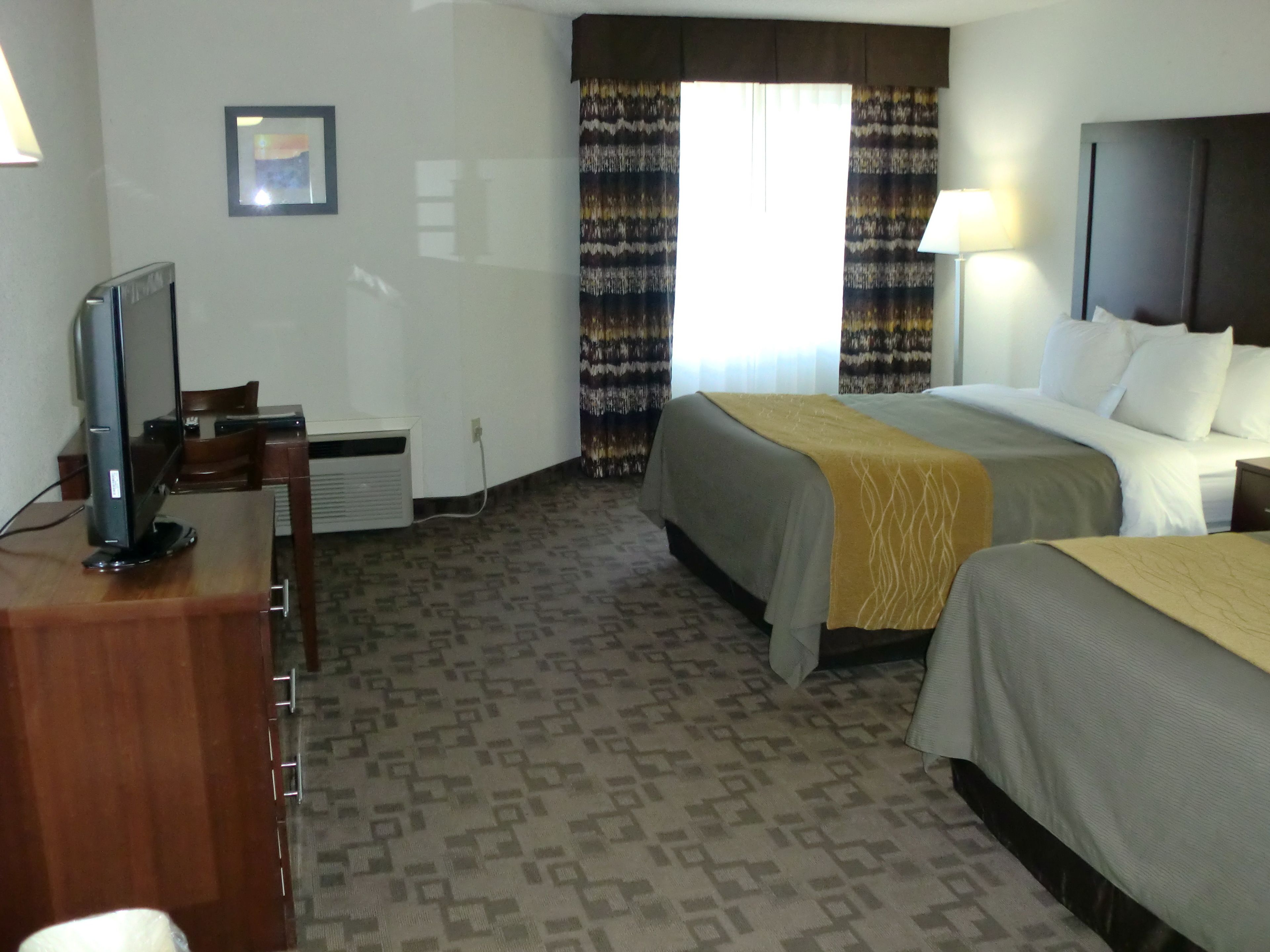 Habitación de hotel con dos camas, un escritorio con computadora, televisor, ventana con cortinas y cabecera.