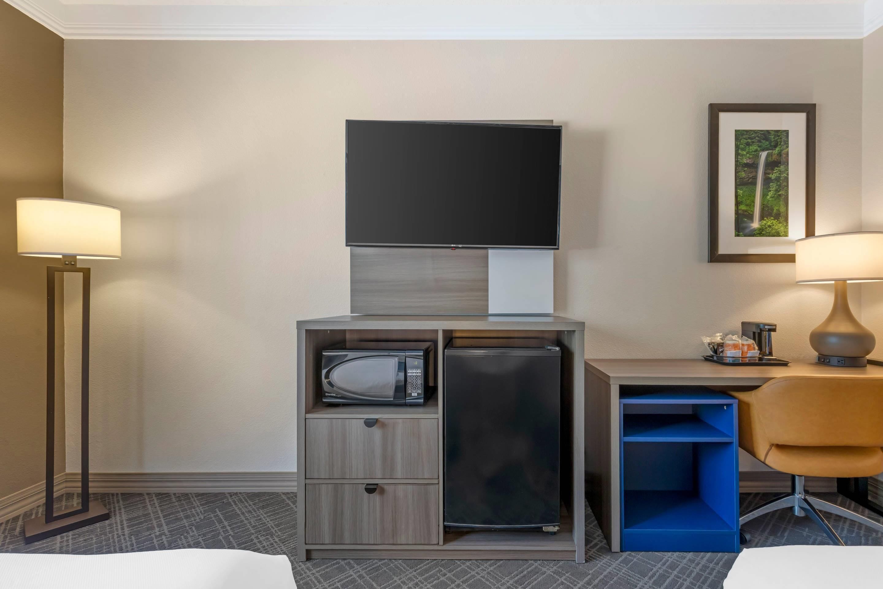 A hotel room with a flat-screen TV, a microwave, a refrigerator, a desk with a chair, and a framed picture on the wall.