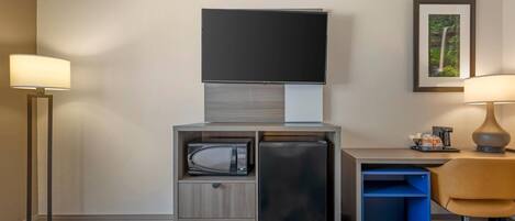 Habitación de hotel con televisor de pantalla plana, microondas, refrigerador, escritorio con silla y un cuadro en la pared.