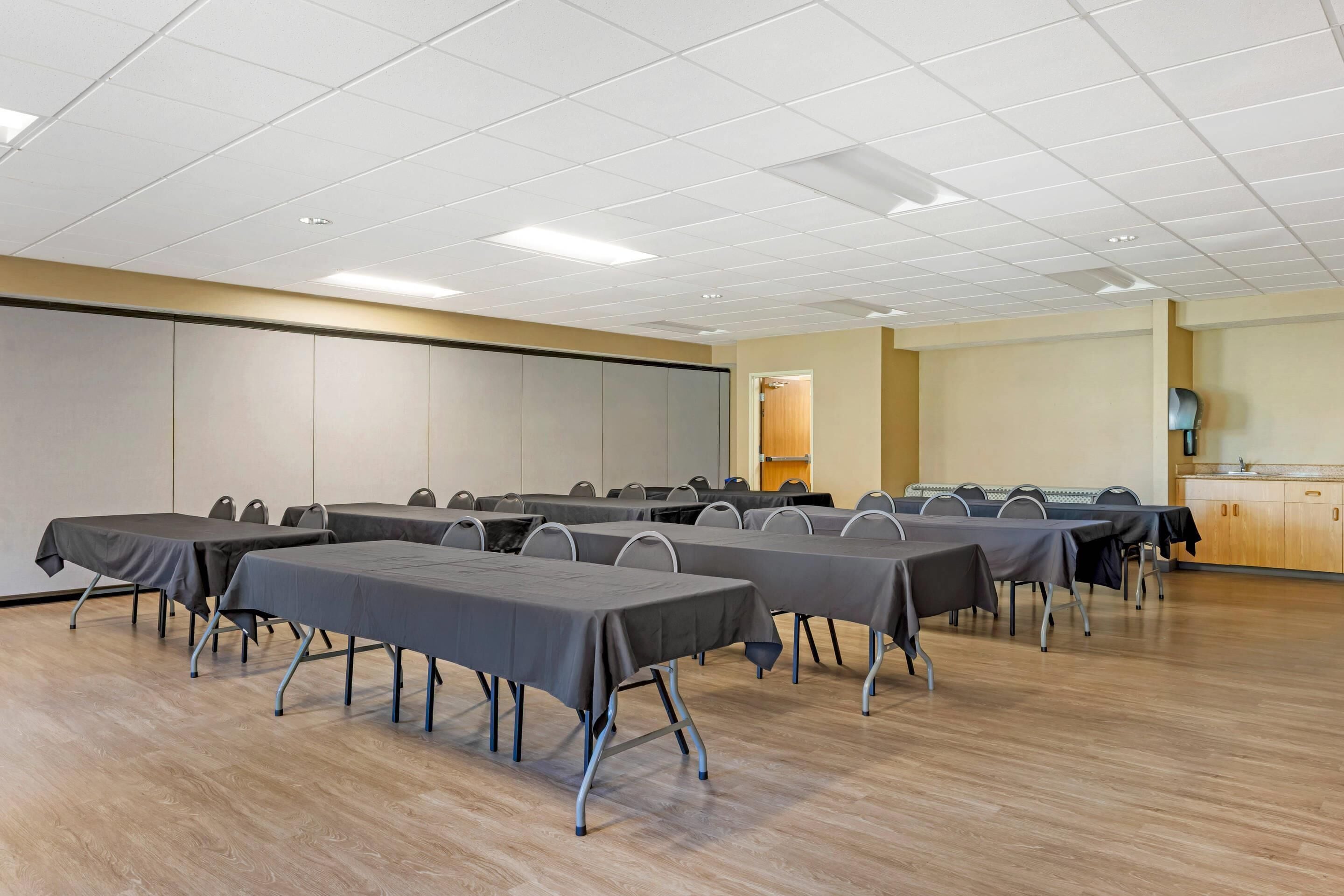 A conference room with multiple tables and chairs, a wooden floor, and a kitchenette in the background.