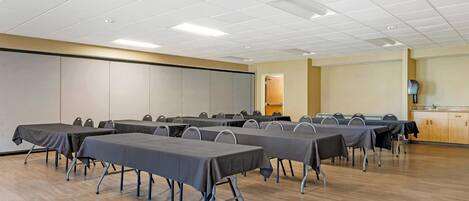 A conference room with multiple tables and chairs, a wooden floor, and a kitchenette in the background.