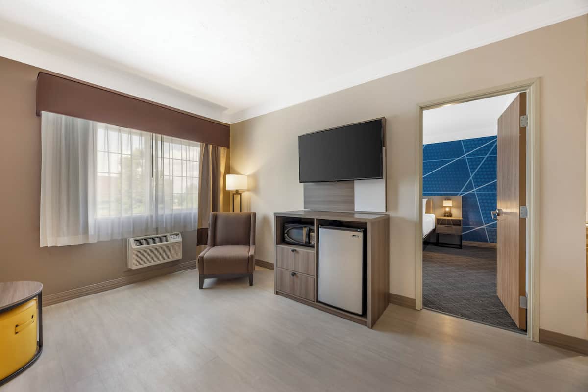 A hotel room with a flat-screen TV mounted above a wooden cabinet, a chair, a desk with a lamp, and a large window with sheer curtains.