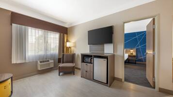 Une chambre d’hôtel équipée d’un téléviseur à écran plat fixé au-dessus d’un meuble en bois, d’une chaise, d’un bureau avec une lampe et d’une grande fenêtre ornée de rideaux légers.