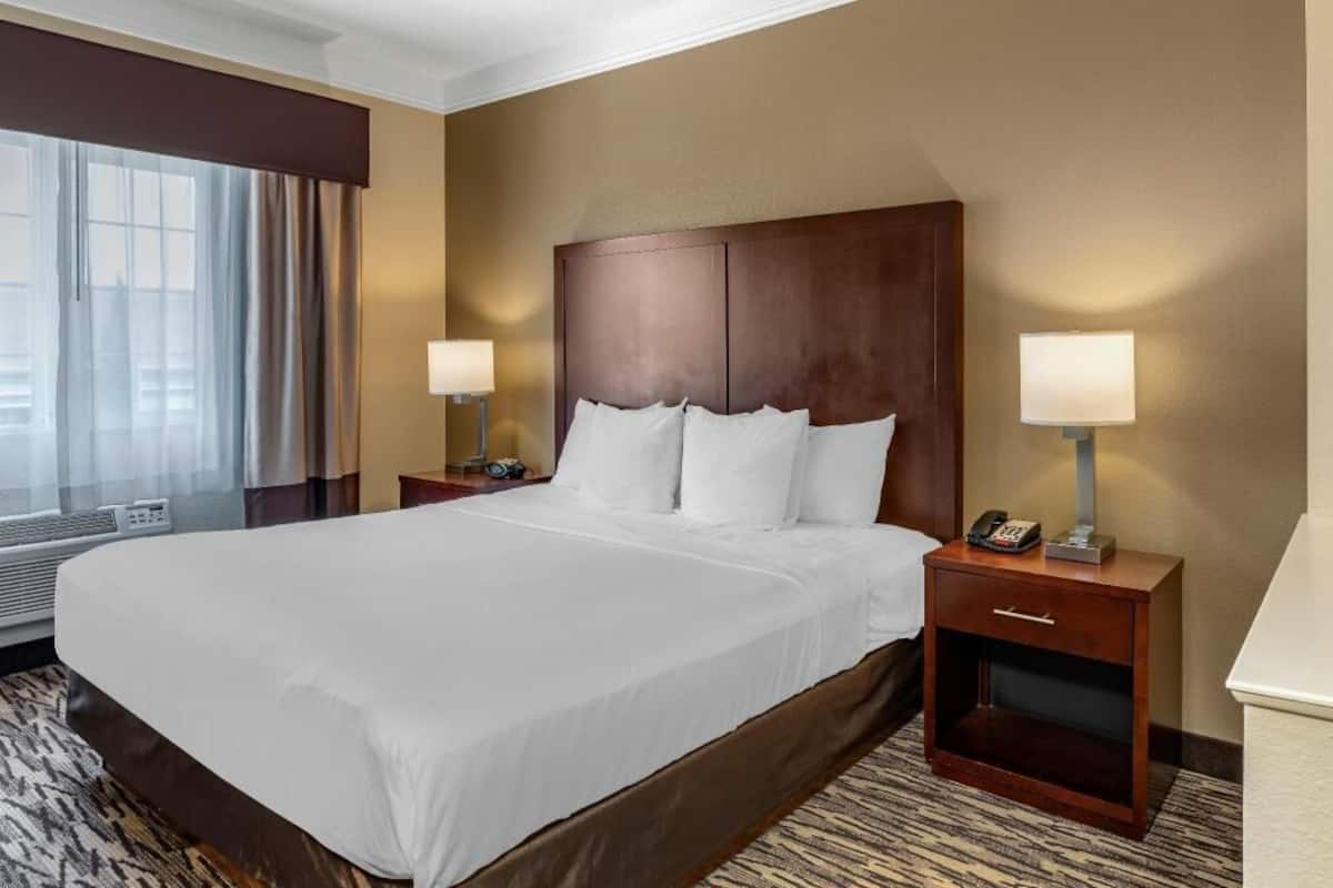 A hotel room with a large bed, two bedside tables with lamps, a window with curtains, and a carpeted floor.
