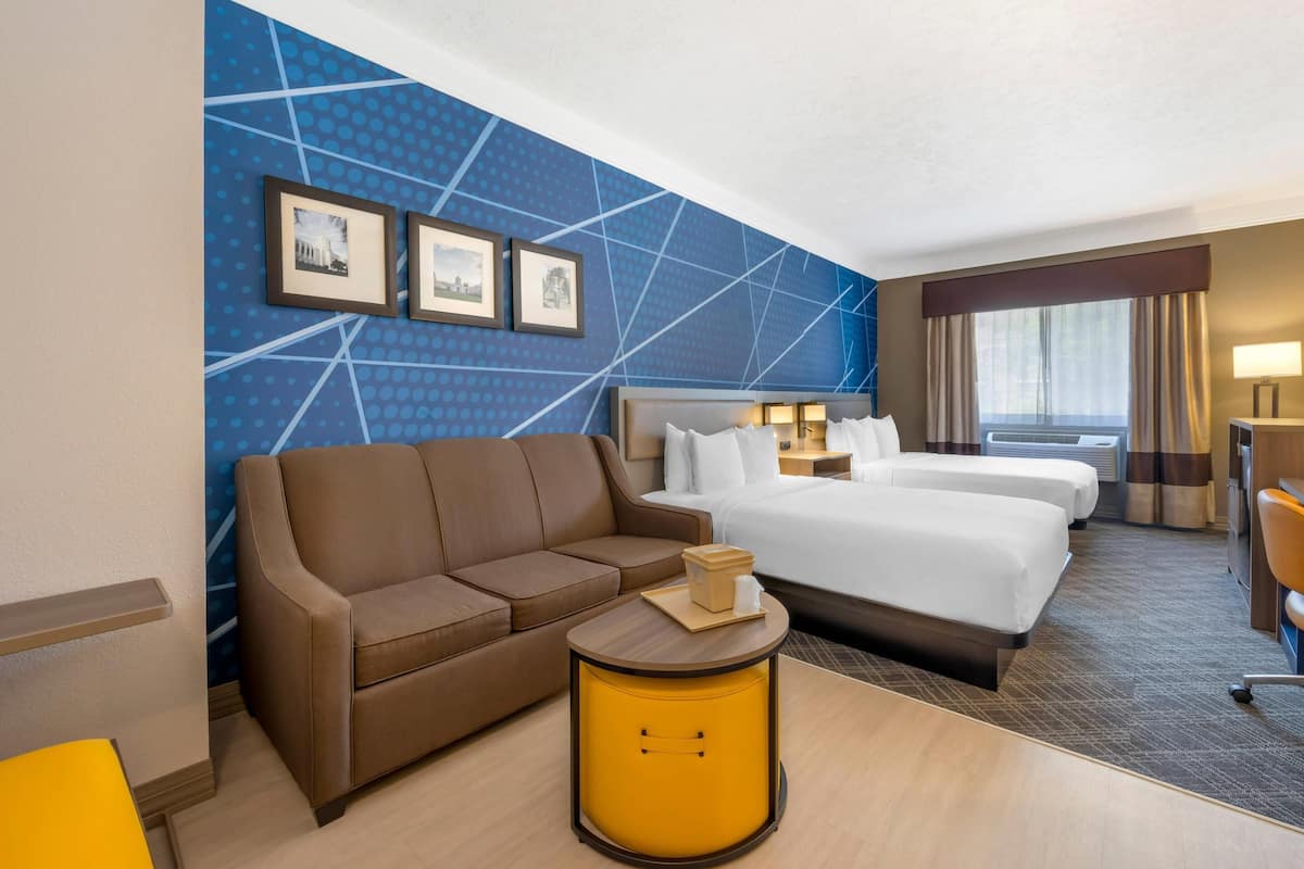 A hotel room with a brown sofa, a bed, a small round yellow side table, and a blue wall with geometric patterns and framed pictures.