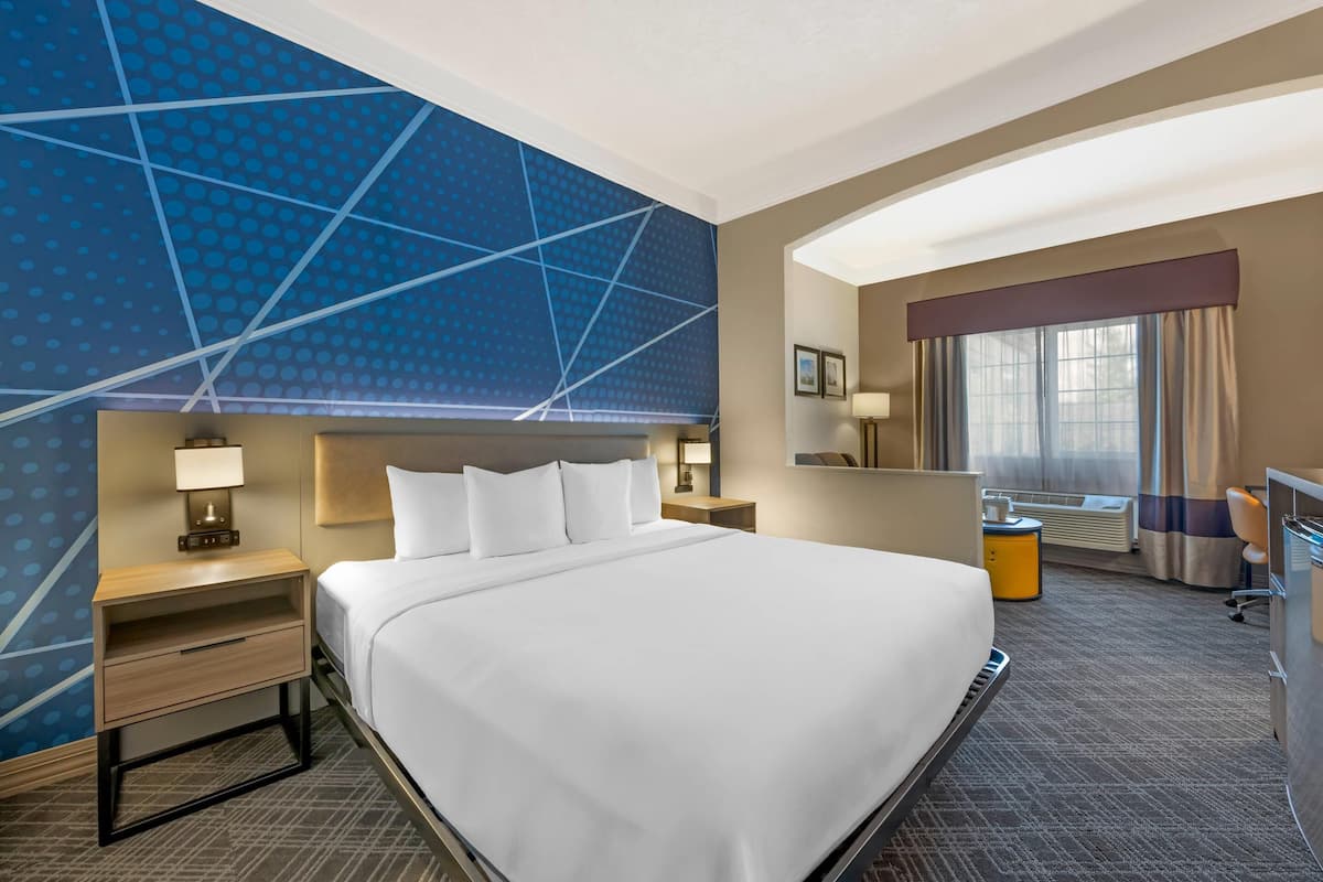 A hotel room with a large bed, a desk, a chair, a desk lamp, a nightstand, a wall-mounted TV, and a window with curtains.