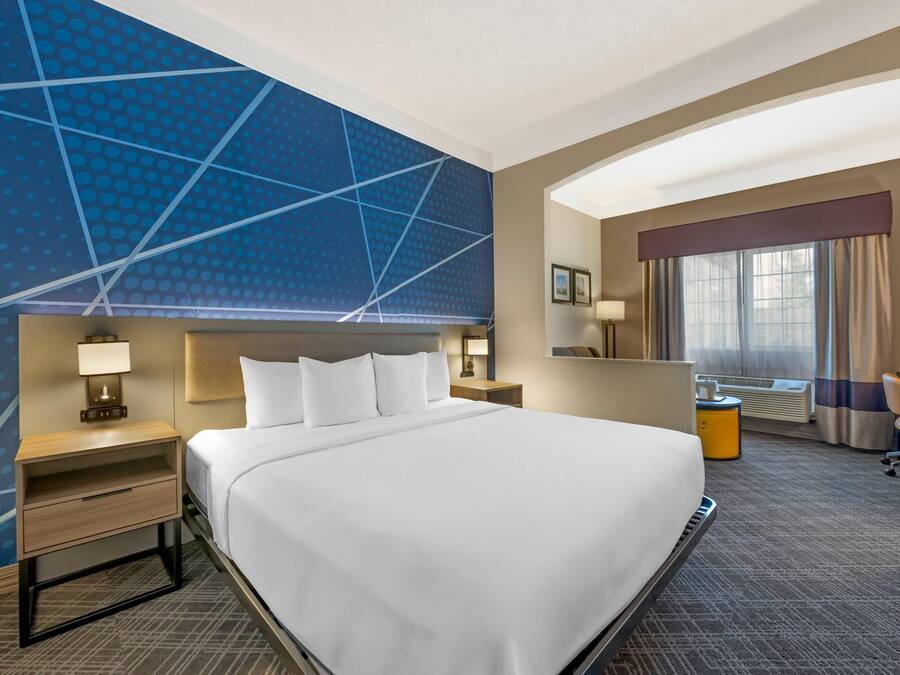 A hotel room with a large bed, a desk, a chair, a desk lamp, a nightstand, a wall-mounted TV, and a window with curtains.