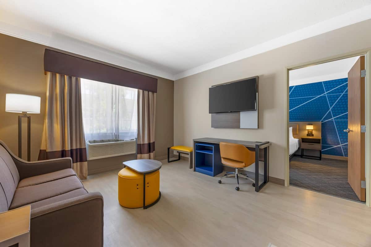A hotel room with a sofa, a small round table, a desk with a chair, a TV, and a window with curtains.
