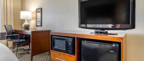 A hotel room with a flat-screen TV, a microwave, a refrigerator, a desk with a chair, and a lamp.
