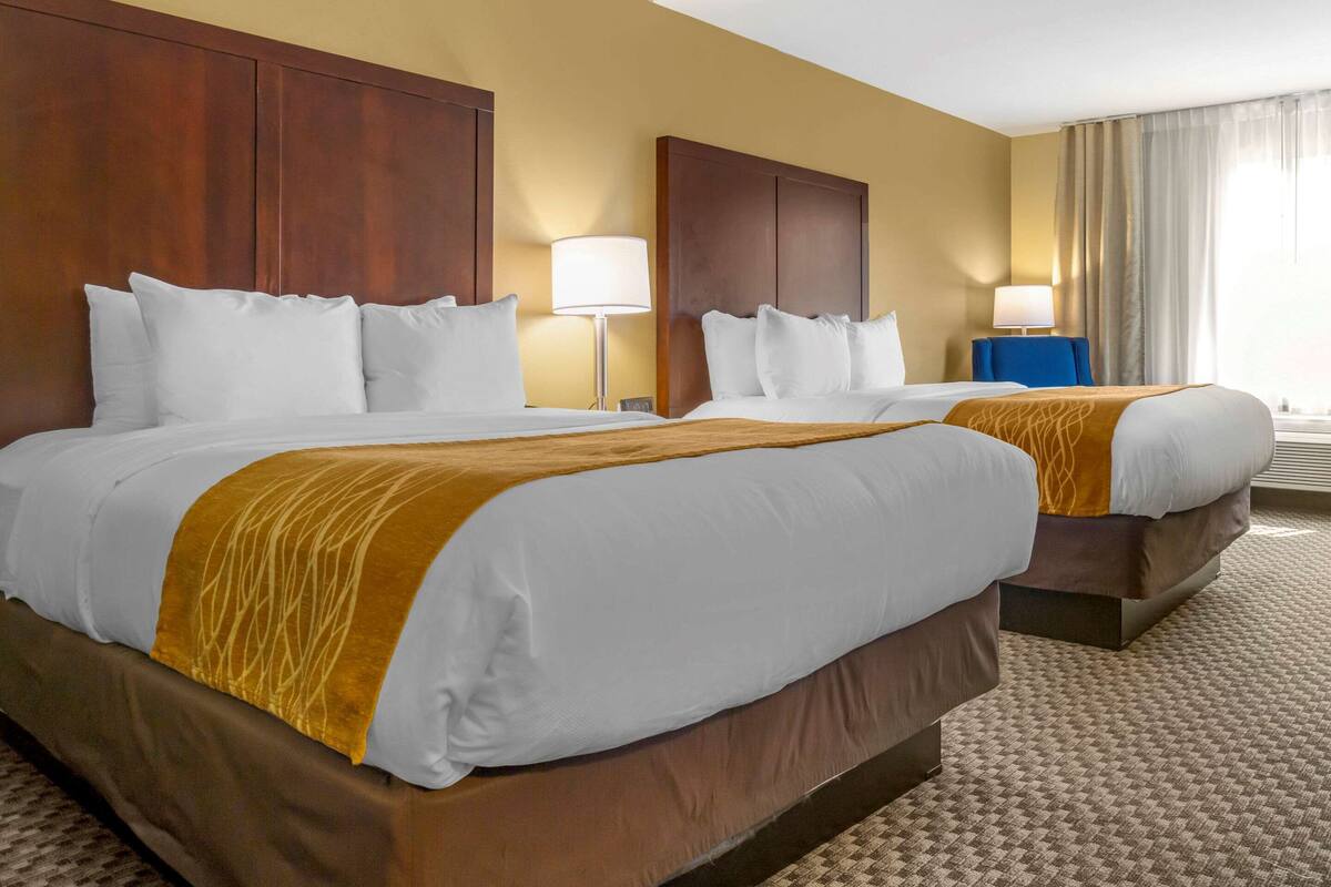 A hotel room with two beds, a wooden headboard, a beige carpet, and a window with curtains.
