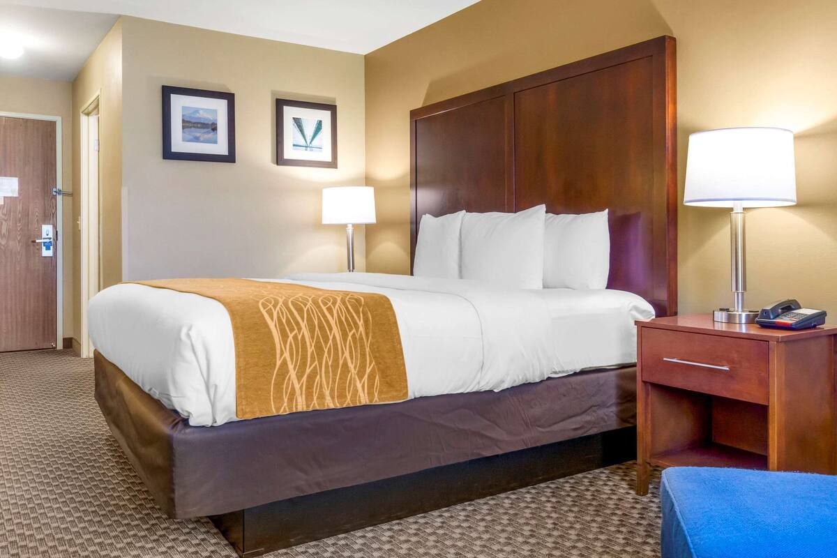 A hotel room with a large bed, a wooden headboard, a bedside table with a lamp, and a small drawer. There are two framed pictures on the wall above the bed.