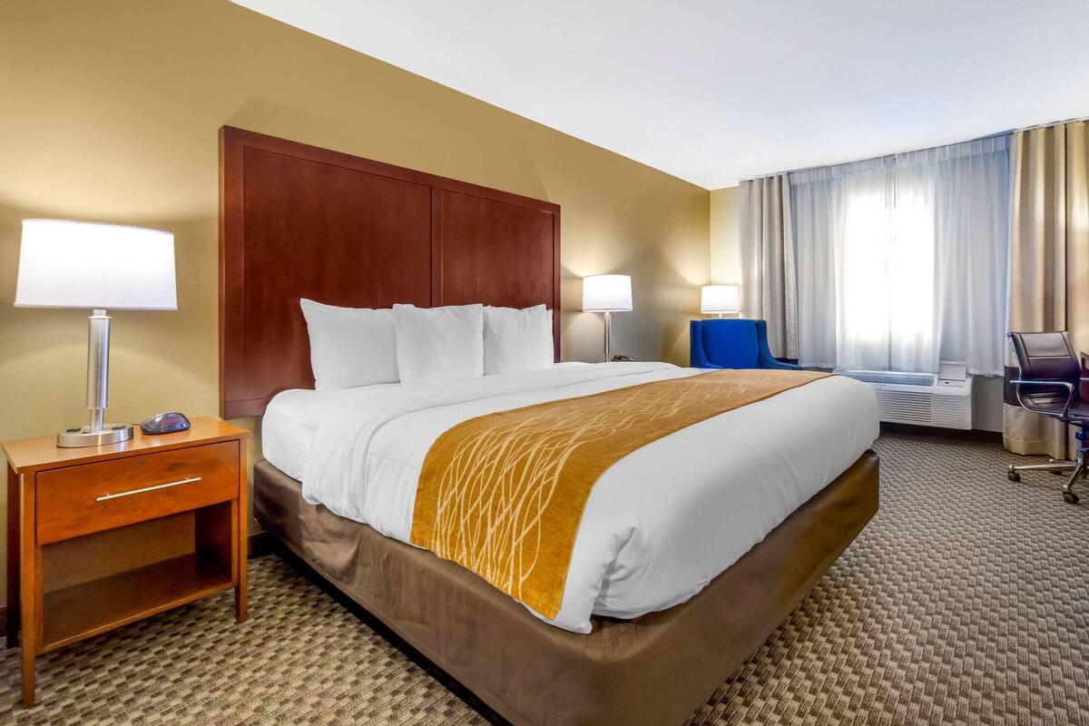 A hotel room with a large bed, wooden headboard, two bedside tables with lamps, a desk with a chair, and a large window with curtains.