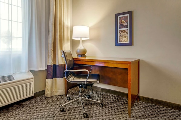 A hotel room with a desk, chair, lamp, air conditioner, and a framed picture on the wall.