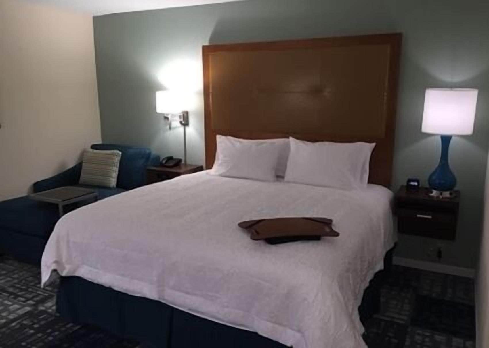 Quarto de hotel com uma cama grande, mesa de cabeceira, luminária e uma cadeira.