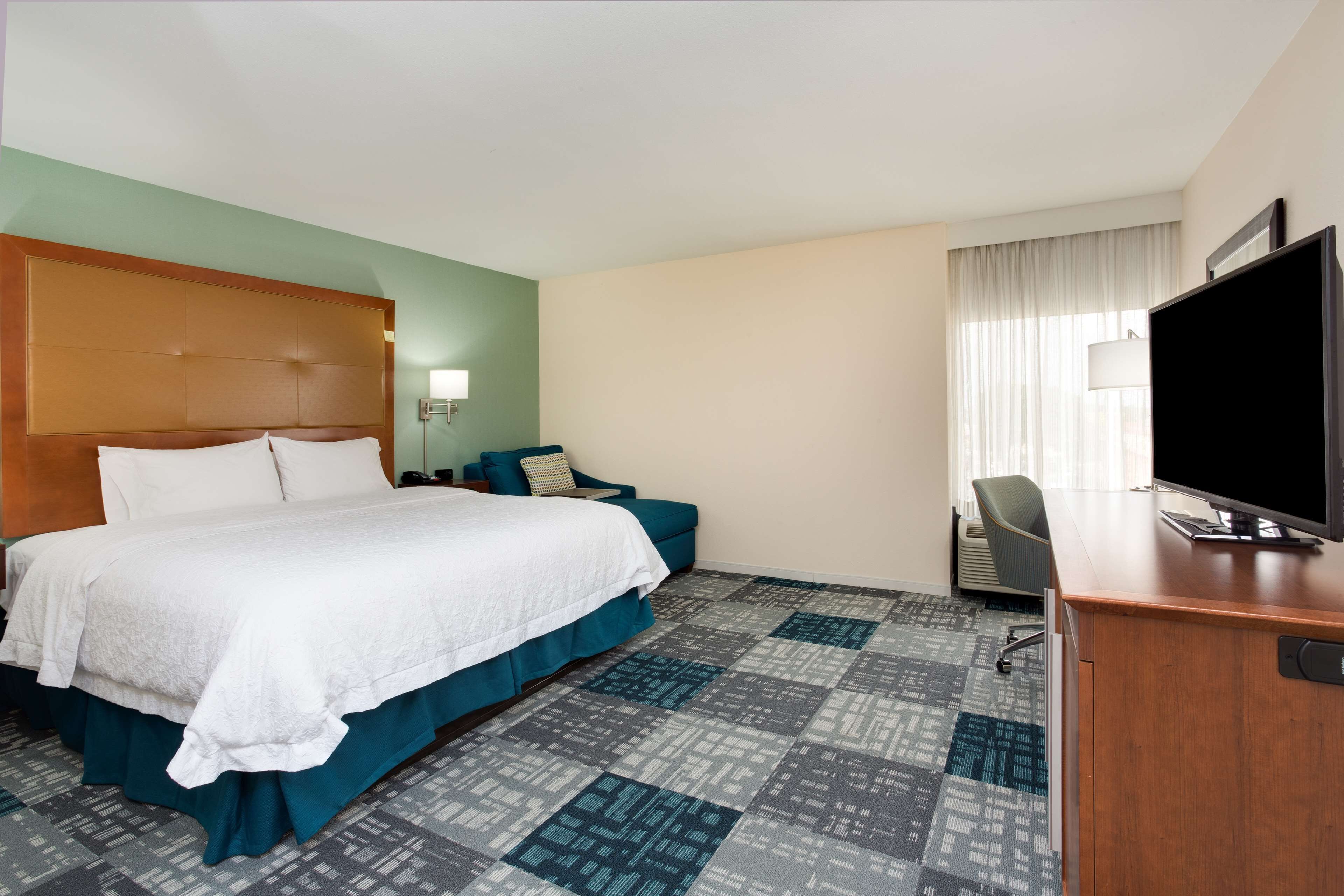 A hotel room with a large bed, a desk with a TV, a chair, and a window with curtains.