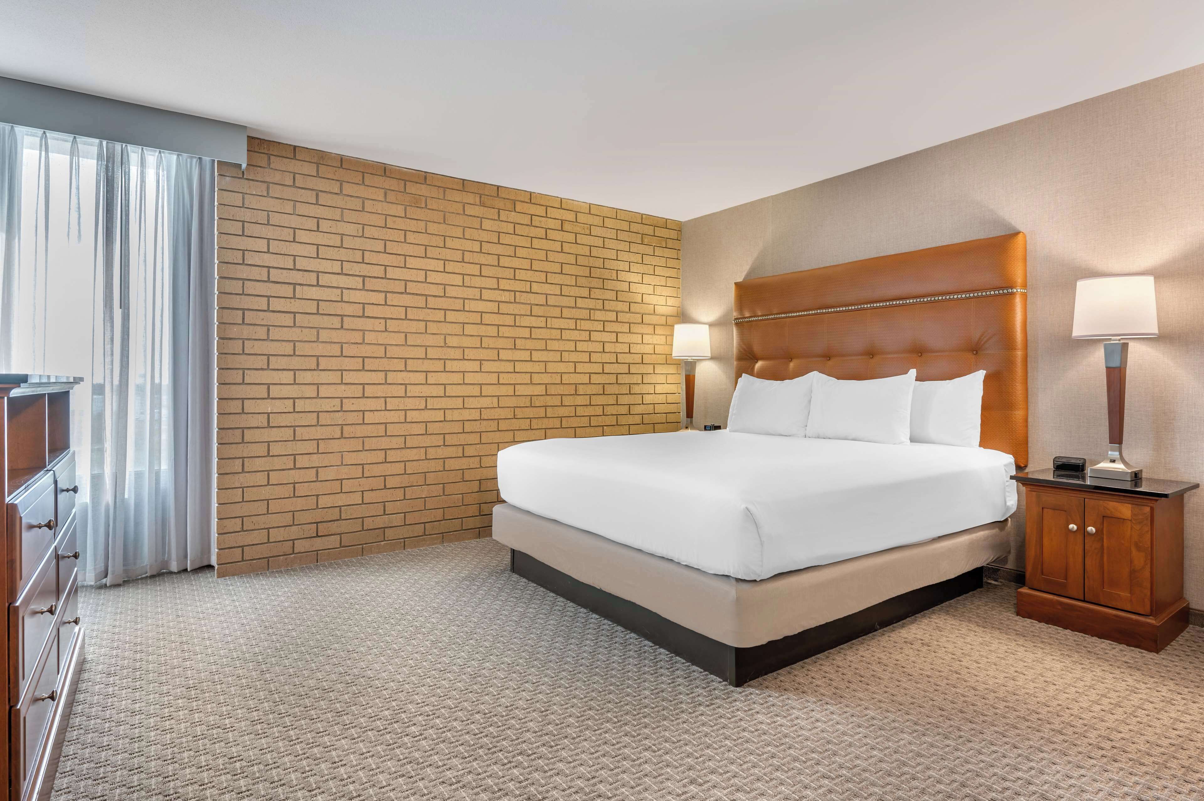A hotel room with a large bed, a wooden nightstand with a lamp, a brick accent wall, and a window with sheer curtains.