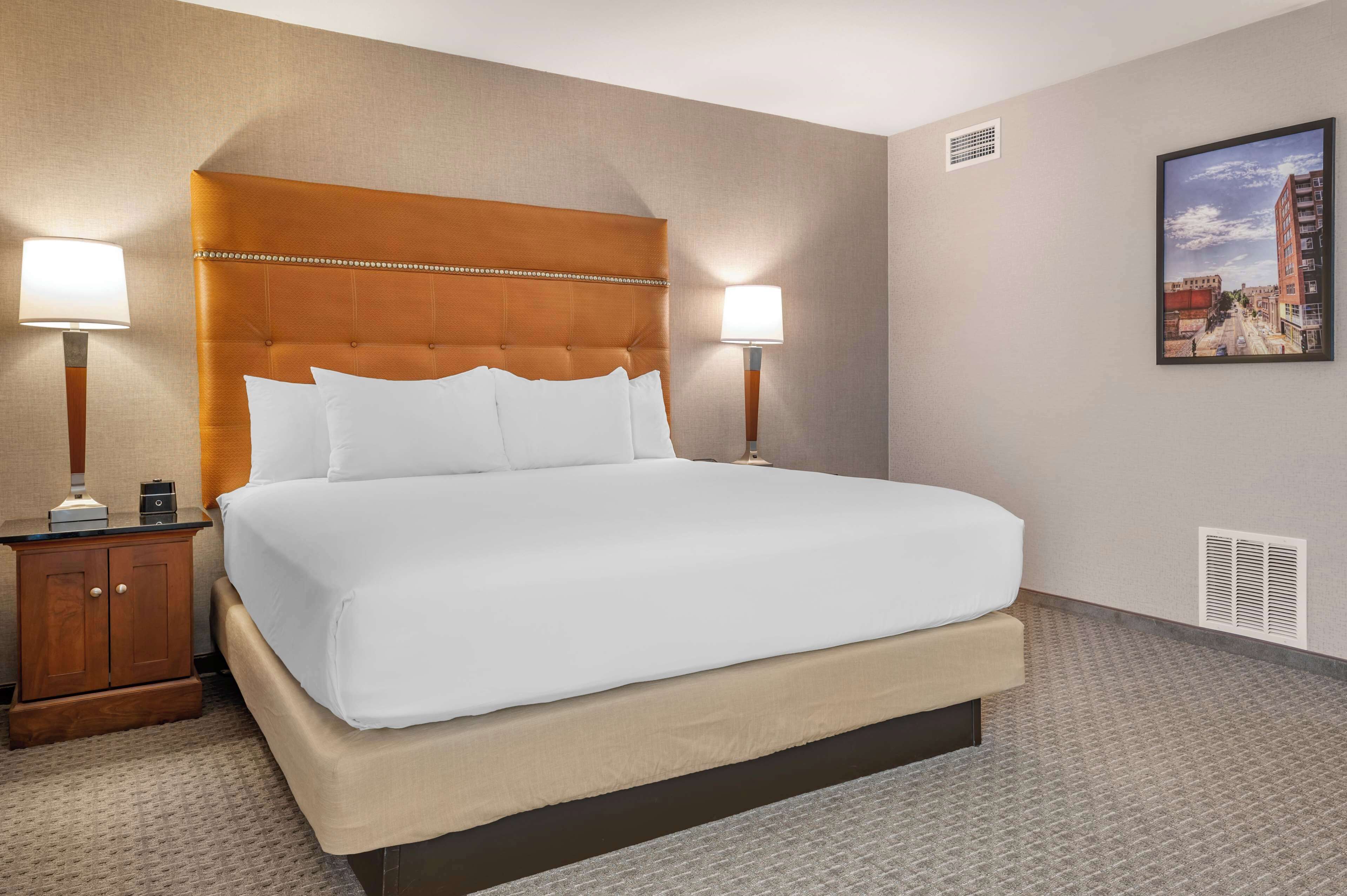 A hotel room with a large bed, two bedside tables, a nightstand, a wall-mounted picture, and a vent.