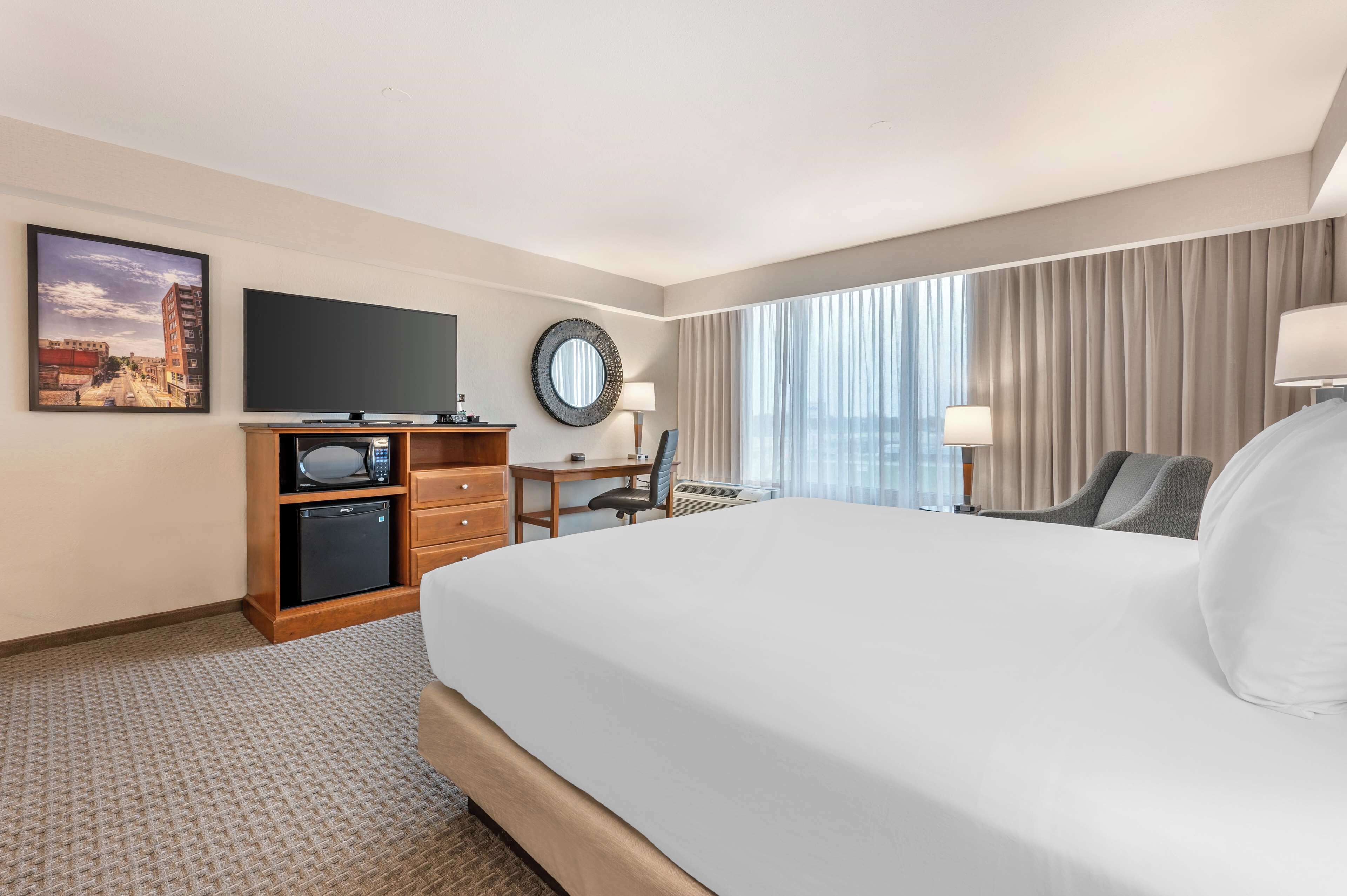 A hotel room with a large bed, a desk with a chair, a television, a small refrigerator, and a window with curtains.