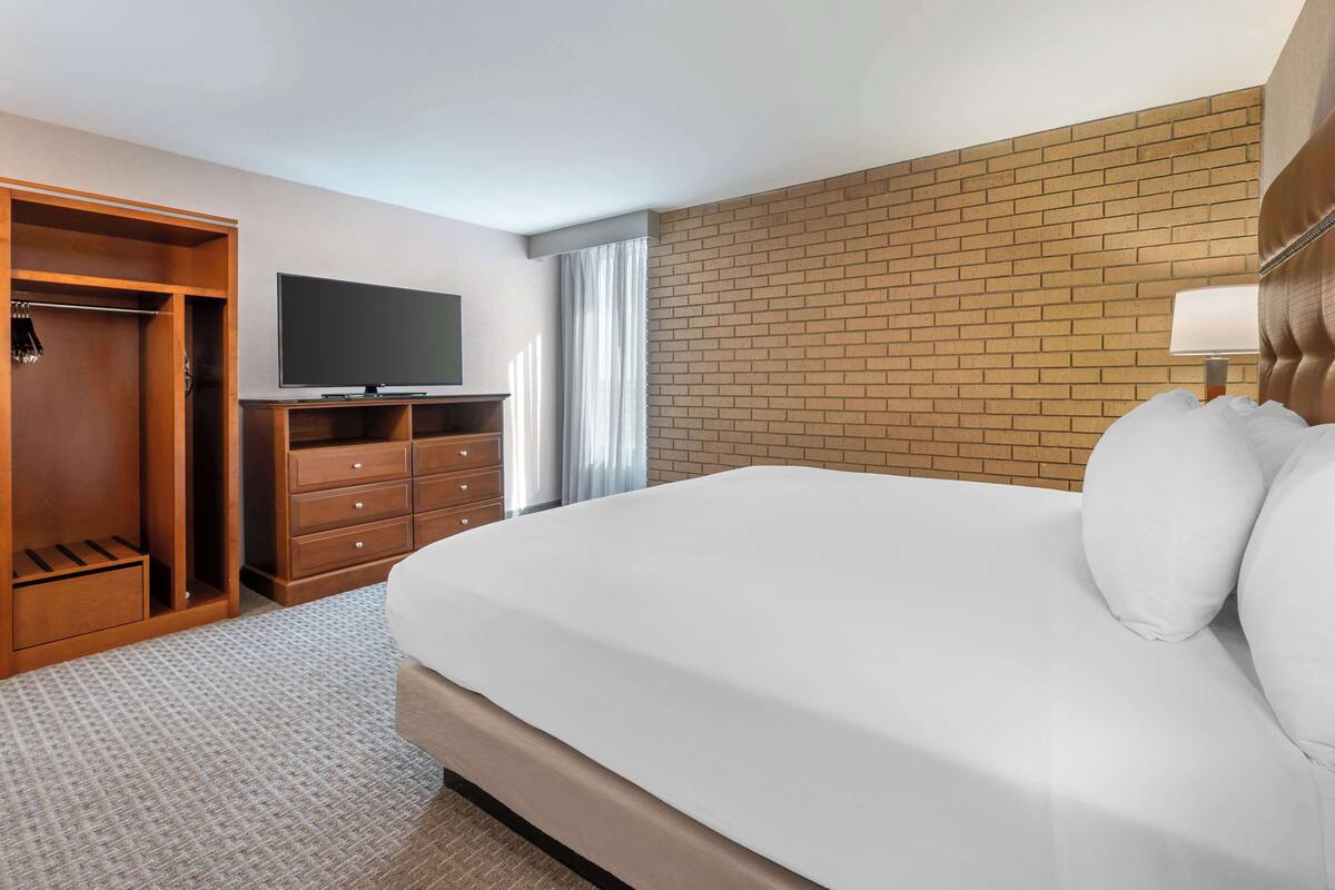 A hotel room with a large bed, a TV on a stand, a wardrobe, and a brick accent wall.