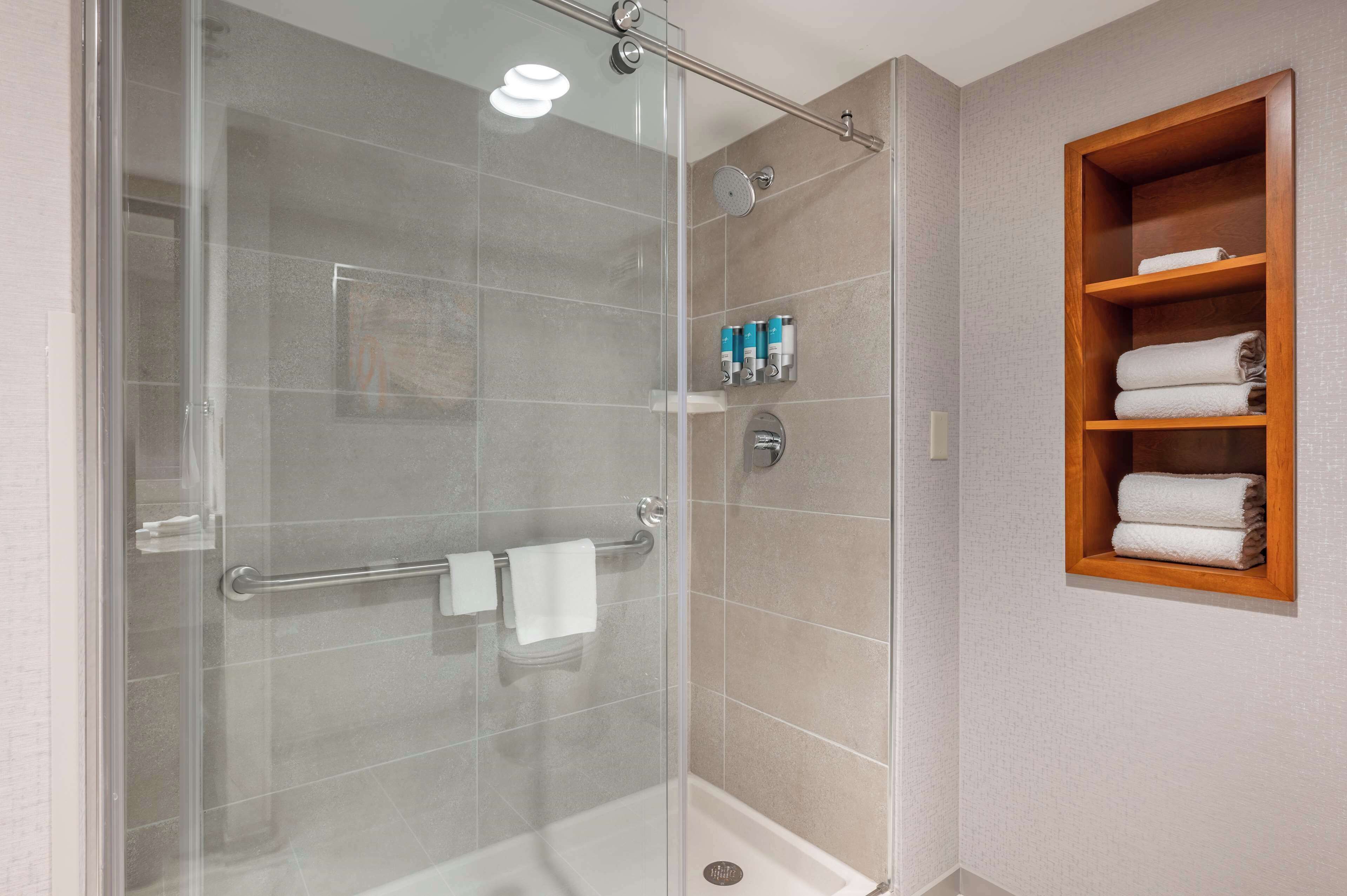 A modern bathroom with a glass-enclosed shower, a wooden towel rack, and a shelf with toiletries.