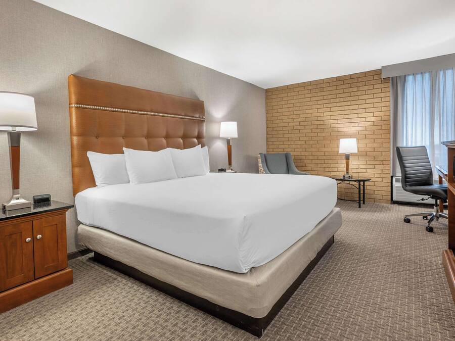 A hotel room with a large bed, two bedside tables with lamps, a desk with a chair, and a brick-patterned wall.