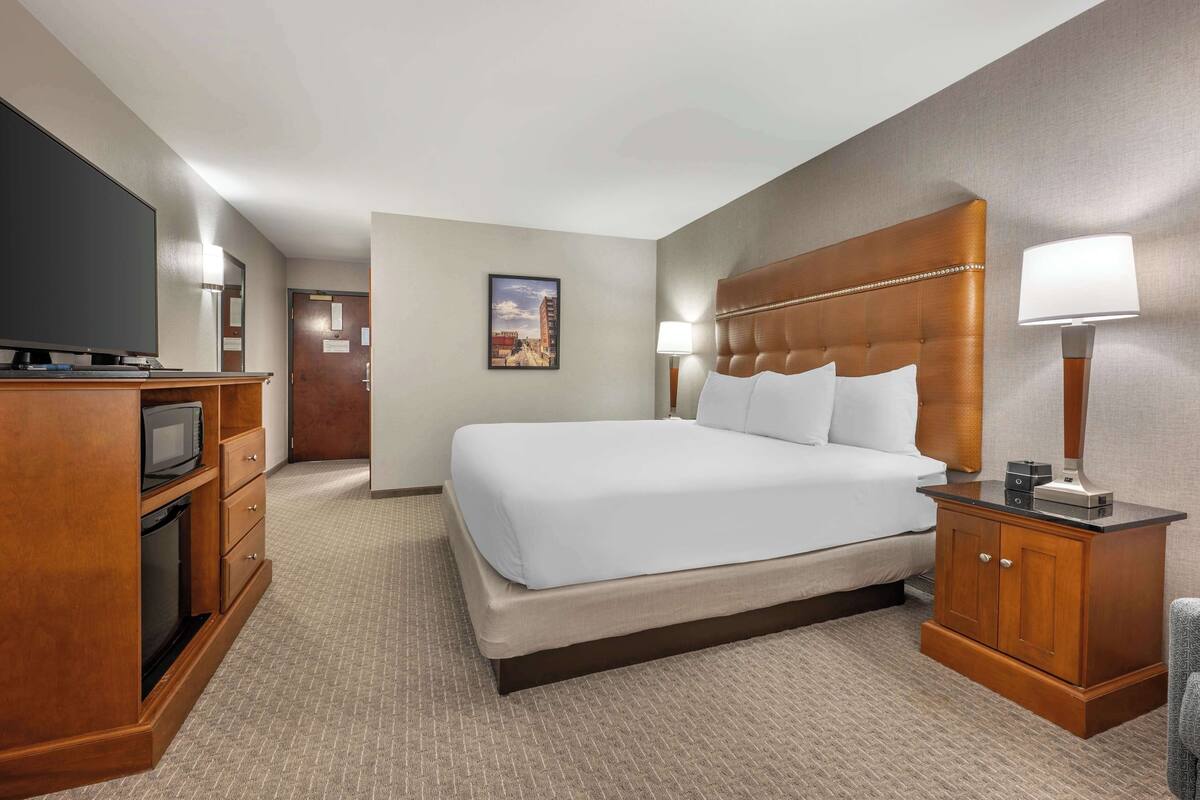 A hotel room with a large bed, wooden headboard, a TV, a desk with a lamp, and a picture on the wall.