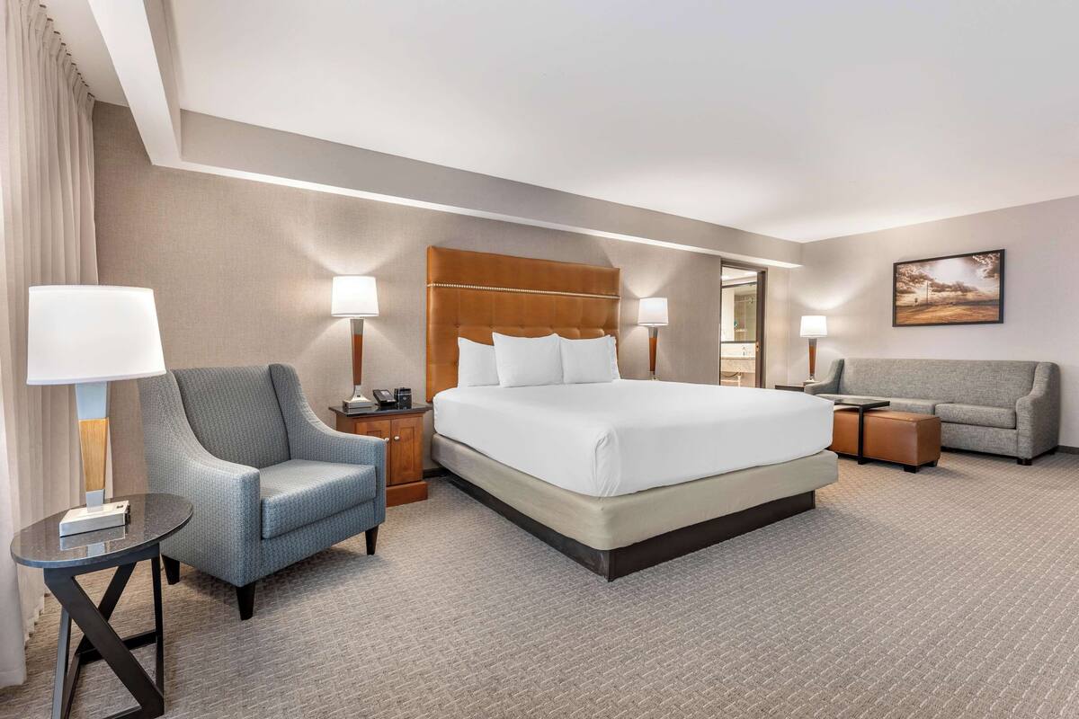 A hotel room with a large bed, a grey armchair, a sofa, and a small table with a lamp.