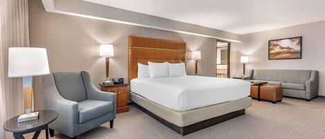A hotel room with a large bed, a grey armchair, a sofa, and a small table with a lamp.
