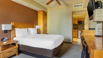 A hotel room with a large bed, wooden headboard, bedside tables, a desk, a ceiling fan, a flat-screen TV, and a kitchenette in the background.