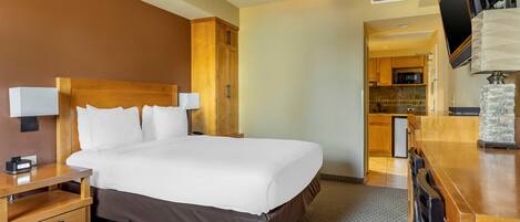 A hotel room with a large bed, wooden headboard, bedside tables, a desk, a ceiling fan, a flat-screen TV, and a kitchenette in the background.